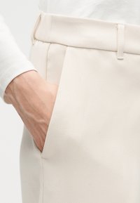 Χέρι μέσα στην τσέπη παντελονιού κρεμ με ραφή και θηλιά ζώνης, φορεμένο με λευκή μπλούζα μακριάς μανίκας.