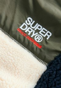Bunda s fleece podšívkou s olivovo zeleným nylonem, smetanovým sherpa materiálem a námořnickými modrými akcenty, s vyšívaným logem "SUPERDRY" v bielej a červenej farbe.
