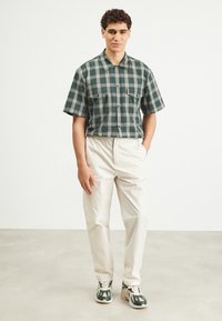 Chemise à manches courtes en tartan vert avec fermeture à boutons, deux poches poitrine, associée à un pantalon beige à taille élastique et des baskets avec des accents verts.