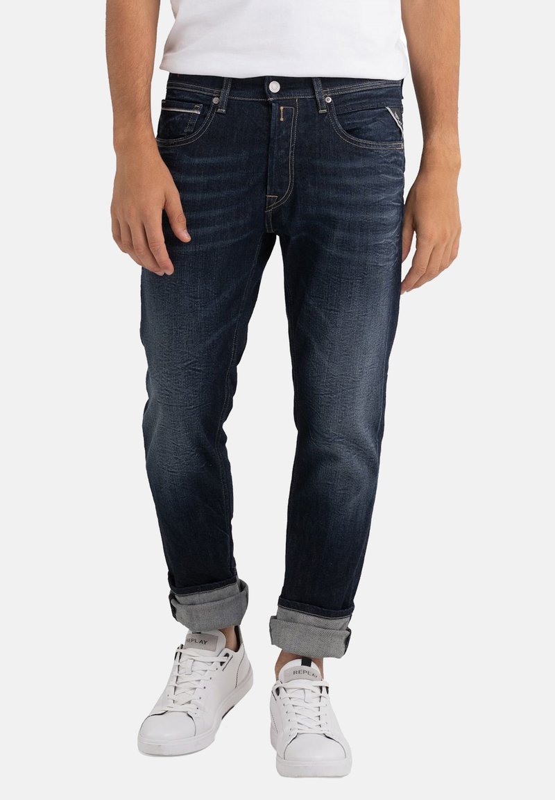 Jeans din denim de culoare albastru închis cu o croială ajustată, având cusături portocalii, margini răsucite și închidere cu nasture în față, asortați cu adidași albi.