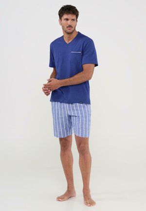 Hombre de pie descalzo con una camiseta azul de manga corta y cuello en V con bolsillo en el pecho, y pantalones cortos a rayas azules y blancas.