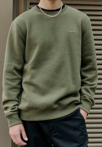 Felpa verde con scollatura a girocollo e polsini a coste. Caratterizzata da un piccolo logo "blend". Tessuto liscio e vestibilità comoda. Pantaloni neri esposti.