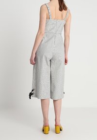 Combinaison rayée avec bretelles réglables, corsage ajusté, jambes larges et raccourcies, et accents de nœuds noirs à l'ourlet. Le modèle porte des chaussures à talons jaunes.