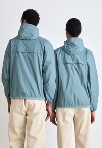 Chaquetas de nylon azul claro con capuchas, con puños elásticos y una pieza de soporte en la parte posterior. Llevadas sobre pantalones color crema, que presentan un corte relajado.