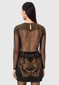 Schwarz Kleid mit transparenten langen Ärmeln, goldenen Perlen auf den Schultern, niedrigem Rückenausschnitt und Paillettenverzierung am Rock, die komplizierte Muster darstellt.