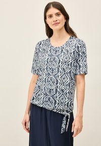 Top blu navy a maniche corte con un motivo geometrico bianco; presenta un collo a barca e un dettaglio con fiocco sul bordo.