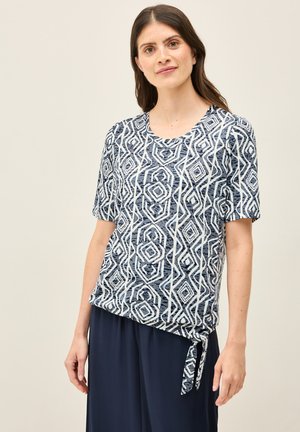 Haut bleu marine à manches courtes avec un motif géométrique blanc ; présente un col rond et un détail noué sur le côté à l'ourlet.
