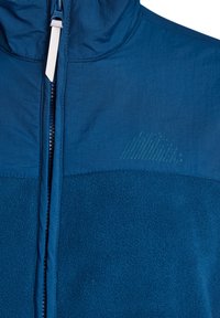 Närbild av en blå fleecejacka med en dragkedja framtill och ett texturerat tygpanel på övre bröstet med subtil logotypdetalj.