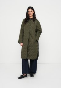 Πράσινο χακί επενδυμένο παλτό με χαλαρή εφαρμογή, που διαθέτει εμπρόσθιο κούμπωμα με κουμπιά, μεγάλες τσέπες και μήκος μέχρι τη μέση. Συνδυασμένο με σκούρο τζιν με φαρδιά μπατζάκια.