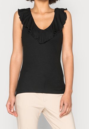 Vrouw draagt een mouwloze zwarte top met ruches aan de V-hals en beige broek, staand tegen een effen lichte achtergrond.