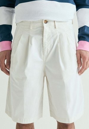 Hombre que lleva pantalones cortos blancos plisados y una camisa de manga larga con bloques de color azul marino, blanco y rosa, de pie con las manos relajadas a los lados.