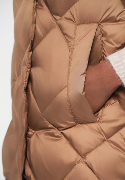 Gilet rembourré brun avec un motif matelassé et une poche latérale. Le matériau semble lisse et brillant, mettant en avant une couleur chaude et neutre.