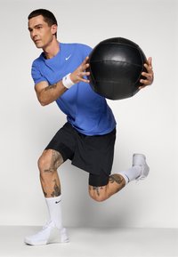 Hombre con camisa azul y pantalones cortos negros haciendo una zancada mientras sostiene una gran pelota medicinal negra, vistiendo zapatillas y calcetines blancos, contra un fondo liso.