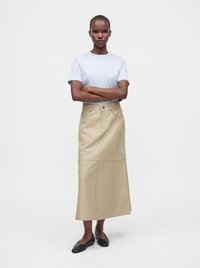 Femme debout, les bras croisés, portant un t-shirt bleu clair, une longue jupe beige et des chaussures plates noires sur un fond blanc uni.
