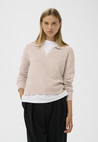 Pulover ribbed roz deschis cu guler în V, purtat peste o cămașă albă, asortat cu pantaloni negri. Textură moale și croi lejer.