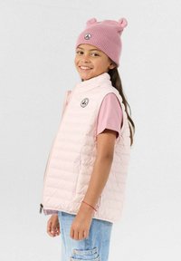 Lichtroze gewatteerd vest met hoge kraag, ritssluiting aan de voorkant en een logo-label. Draagt over een lichtroze T-shirt, gecombineerd met spijkerbroek en een roze muts.