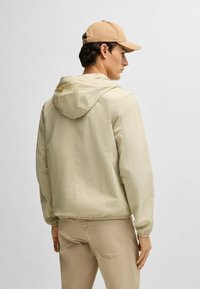 Light beige, lightweight jacket with a hood and elastic cuffs, paired with matching beige pants. The setting is neutral, unobtrusive background.