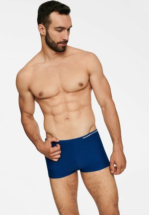 Navy blue swim trunks made of stretchy material, featuring a snug fit, a flat waistband with branding, and a smooth texture.