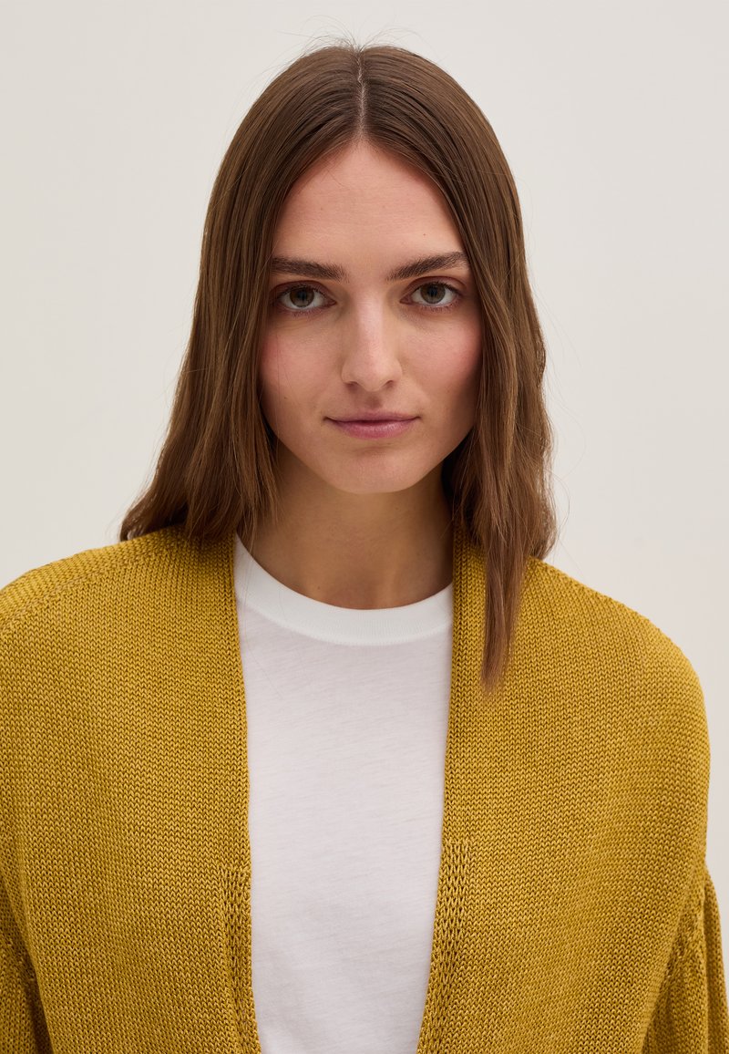 Joven mujer con cabello castaño liso, llevando un cárdigan de punto amarillo mostaza sobre una camisa blanca, fondo neutro.