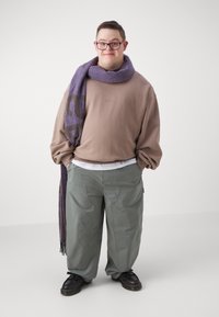 Uomo con occhiali che indossa una felpa beige, pantaloni grigi larghi, scarpe nere e una lunga sciarpa viola su uno sfondo bianco semplice.