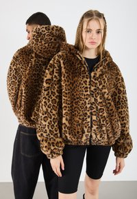 Două modele îmbrăcate cu jachete din blană sintetic�ă cu imprimeu de leopard maro, una cu fața spre cameră purtând pantaloni scurți negri, cealaltă fiind cu spatele întors purtând pantaloni negri.