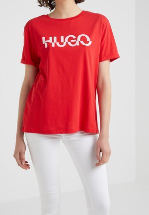 Personne portant un T-shirt rouge aux manches retroussées avec le texte blanc « HUGO », associé à un pantalon blanc ajusté sur un fond uni.
