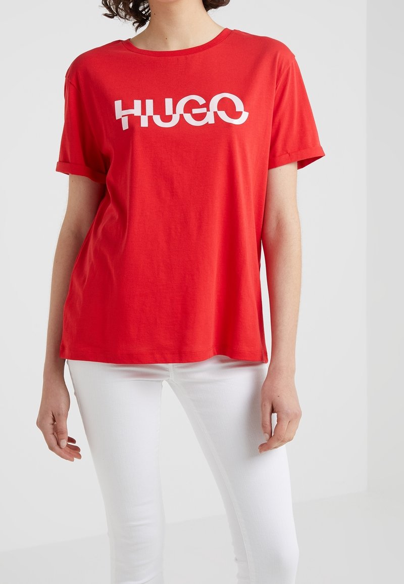 Persona con una camiseta roja de mangas remangadas y texto blanco "HUGO", combinada con pantalones blancos ajustados sobre un fondo liso.