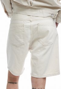 Shorts de algodón beige con corte recto, que cuentan con dos bolsillos traseros, detalles de costura visibles y un dobladillo limpio a la altura de la rodilla.