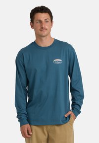 Mężczyzna z krótkimi brązowymi włosami i wąsem, ubrany w turkusową koszulę z długim rękawem z małym logo Billabong oraz w beżowe spodnie, z ręką w kieszeni.