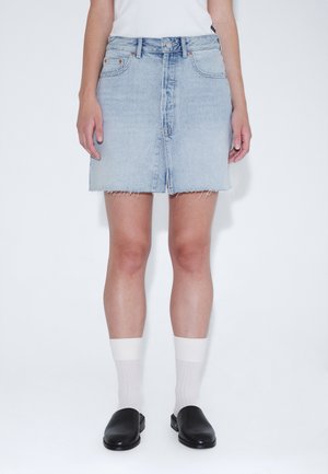 Person iført en lyseblå denimnederdel med frynset kant, hvide sokker, sorte slip-on sko og en hvid top, stående mod en ensfarvet hvid baggrund.