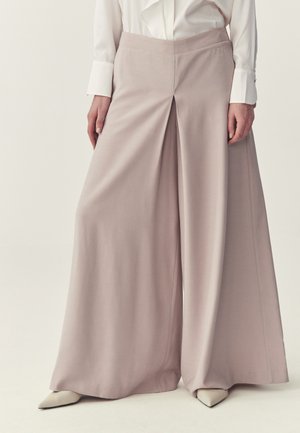 Kobieta w szerokich, jasnobeżowych spodniach z przodu plisowanych, do tego białe szpiczaste buty i biała koszula z długim rękawem.