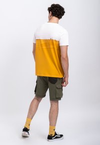 T-shirt met korte mouwen, wit aan de bovenkant en een levendig gele onderzijde. Gecombineerd met olijfgroene shorts en zwarte sneakers met gele sokken.