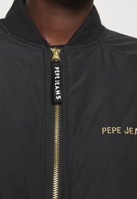 Gros plan d'une veste noire avec une fermeture éclair dorée et "PEPE JEANS" brodé et imprimé près de la fermeture éclair au niveau de la poitrine.