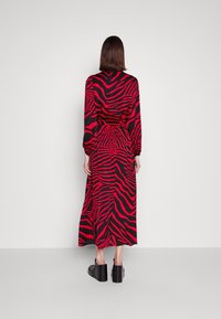 Robe longue à imprimé zèbre noir et rouge, manches longues, taille cintrée, tissu fluide. Chaussures à talons carrés. Fond uni.