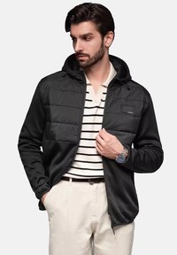 Giacca nera con zip e cappuccio, che unisce tessuto liscio e maniche textured, con taschino sul petto e dettagli con cordino.