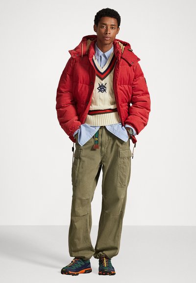 Geacă puf galbenă, pulover crem cu tricotaj în vrăjitorie și decolteu în V, cămașă butonată bleu deschis, pantaloni cargo oliv și pantofi de drumeție multicolori.