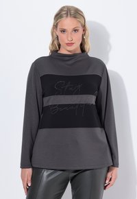 Haut à manches longues en gris foncé et noir, avec un col montant et des rayures horizontales, orné de texte en sequins disant "Restez Belle" sur la poitrine.