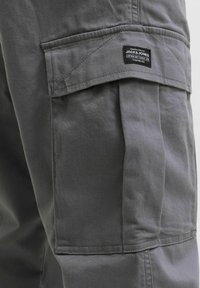 Grå cargo-bukselomme med klaff og svart, rektangulært Jack & Jones-merkelapp sydd midt øverst på lomma.