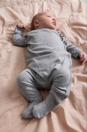 Grå langermet bodysuit på en baby, med trykknapper og myk stoff, liggende på en rynkete beige bakgrunn.