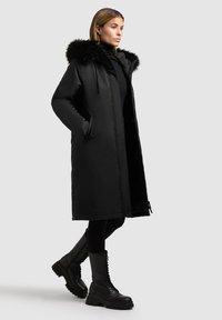 Sort lang frakke med en faux fur hætte, der har en lynlås lukning, to sidelommer og en glat, blank tekstur. Båret med sorte kampstøvler.