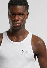 Hvit ribbet tank topp med lett tekstur og svart signaturlogo. Har rund hals og brede ermehull.