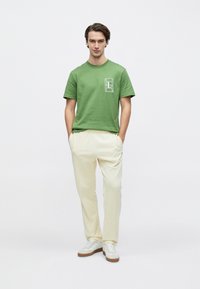 Πράσινο βαμβακερό μπλουζάκι με ορθογώνιο λευκό λογότυπο στο αριστερό στήθος, συνδυασμένο με κρεμ χαλαρά παντελόνια και λευκά αθλητικά παπούτσια.