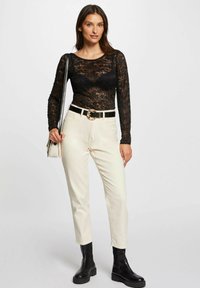 Top lungo a maniche lunghe in pizzo nero, pantaloni a sigaretta beige, cintura nera con fibbia rotonda e stivaletti neri. Modella che tiene una piccola borsa a tracolla.