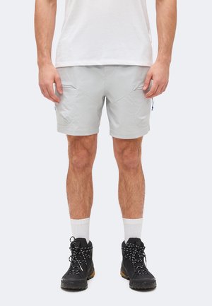 Férfi világosszürke rövidnadrágban, fehér bokazokniban, fekete túracipőben és fehér pólóban, egyszínű háttér előtt áll.