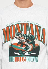 Sweat-shirt blanc avec un grand texte orange et vert "Montana" et "The Big Country", mettant en avant un poisson sautant et un paysage de montagnes.