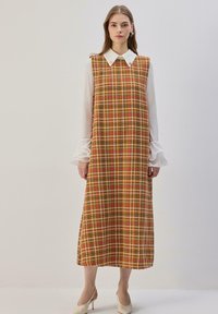 Robe à carreaux marron avec une chemise blanche à col en dessous. Caractérisée par un design sans manches, une longueur jusqu'aux genoux et une texture fluide. Talons blancs.