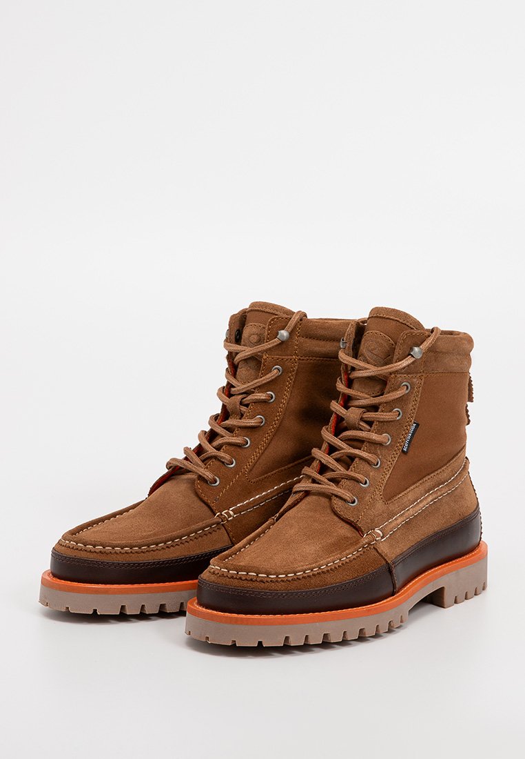 Braune Wildlederschuhe mit einem gepolsterten Kragen, cremefarbener Naht und einem kontrastierenden dunkelbraunen Lederelement. Dicke Gummisohle mit orangefarbenen Details.