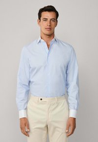 Uomo che indossa una camicia azzurro chiaro con polsini bianchi e pantaloni beige, in piedi contro uno sfondo grigio chiaro uniforme.