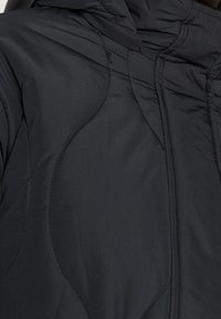 Černá prošívaná bunda s oblými stehy, kapucí a jemnou texturou. Materiál působí lehce a odolně vůči povětrnostním vlivům.