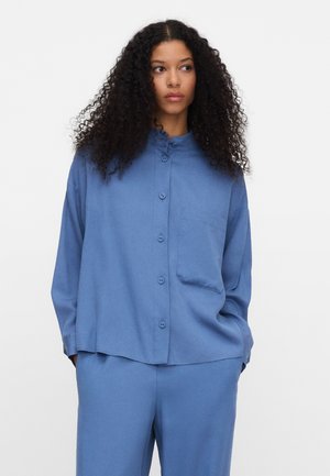 Vrouw met lang krullend haar, gekleed in een loszittend blauw overhemd met knopen en bijpassende blauwe broek, tegen een effen witte achtergrond.